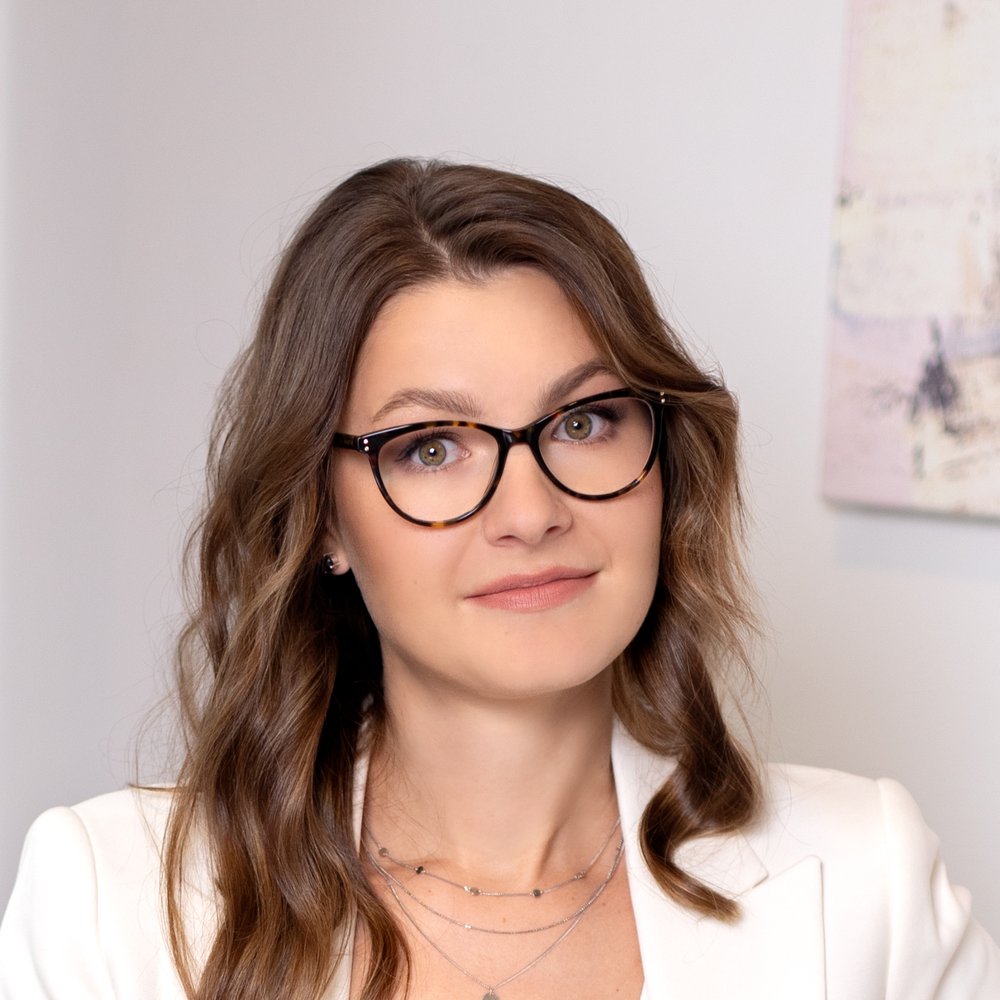 Natalia in a cream suit and glasses, sitting on a grey sofa
